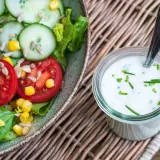Frischer Salat mit Tomaten, Gurken und Mais neben einem Glas mit selbstgemachtem Joghurt-Dressing, garniert mit Schnittlauch.