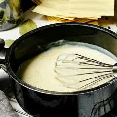 Cremige Béchamelsauce wird in einem Topf mit einem Schneebesen verrührt. Daneben liegen Lasagneplatten, Lorbeerblätter und Gewürze.