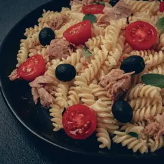Ein köstlicher italienischer Nudelsalat mit Fusilli, Thunfisch, Kirschtomaten und schwarzen Oliven auf einem schwarzen Teller.