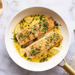 Zwei saftige Lachsfilets in einer Pfanne mit Zitronenscheiben, Kapern und Kräutern. Ein köstlicher Lachs mit Mangold-Aroma.