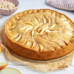Ein goldbrauner, saftiger apfelkuchen ohne ei, dekoriert mit Apfelscheiben. Daneben liegen frische Äpfel und eine Schüssel Haferflocken.