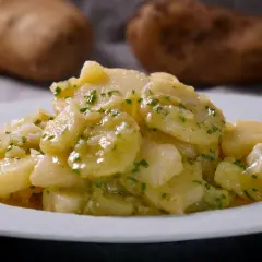 Ein Teller mit klassischem deutschen Kartoffelsalat, garniert mit frischen Kräutern. Im Hintergrund sind rohe Kartoffeln zu sehen.