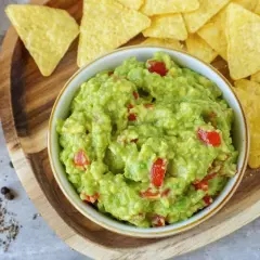 Frisches Guacamole-Rezept mit Tomatenstückchen, serviert mit Tortilla-Chips auf einem Holzbrett.