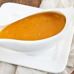 Eine köstliche, goldbraune Sauce mit Fond aus dem Glas, serviert in einer weißen Sauciere auf einem Leinentuch.