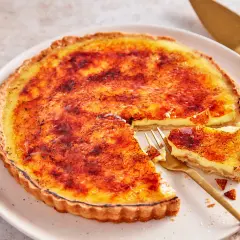Eine cremige Crème brûlée Tarte mit karamellisierter Zuckerkruste, ein Stück wurde mit einer goldenen Gabel entnommen.