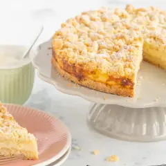 Ein saftiger Streuselkuchen mit Puddingfüllung, bestäubt mit Puderzucker, wartet auf Genießer. Daneben eine Tasse Cappuccino.