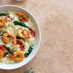 Ein Teller mit Orzo-Nudeln, Garnelen, Spinat und Tomaten, garniert mit Pfeffer. Dazu eine Zitronenspalte.