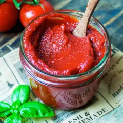 Selbstgemachter Ketchup aus Tomatenmark, mit frischem Basilikum und reifen Tomaten im Hintergrund.