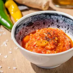 Schüssel mit würzigem Tomaten-Chutney, garniert mit Senfkörnern. Daneben liegen grüne und gelbe Chilis, perfekt für ein Tomaten-Chutney-Rezept.