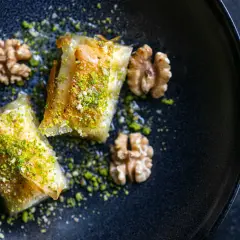 Zwei goldbraune Baklava-Stücke, bestreut mit gehackten Pistazien und Nüssen, auf einem dunklen Teller. Ein köstliches Baklava Rezept.