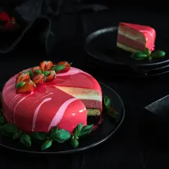 Eine Mousse Torte mit glänzendem Spiegelglasur, dekoriert mit Erdbeeren und Basilikum. Ein Stück ist auf einem Teller angerichtet.