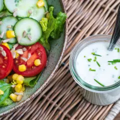 Frischer Salat mit Tomaten, Gurken und Mais neben einem Glas mit selbstgemachtem Joghurt-Dressing, garniert mit Schnittlauch.