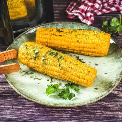 Zwei goldgelbe Maiskolben, frisch aus dem Airfryer, mit Kräutern bestreut, liegen auf einem Teller. Daneben Salz- und Pfefferstreuer.