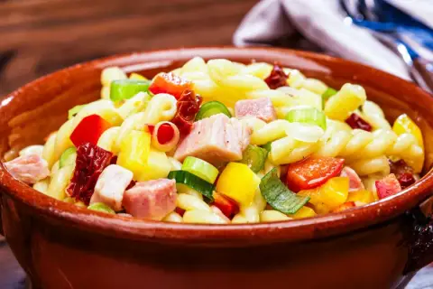 Leckerer nudelsalat ohne mayonnaise mit bunten Paprikawürfeln, Schinken und Frühlingszwiebeln in einer Tonschüssel.