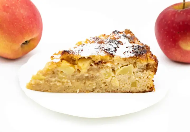 Ein Stück Apfelkuchen ohne Zucker mit Quark, bestäubt mit Puderzucker. Zwei Äpfel liegen daneben.