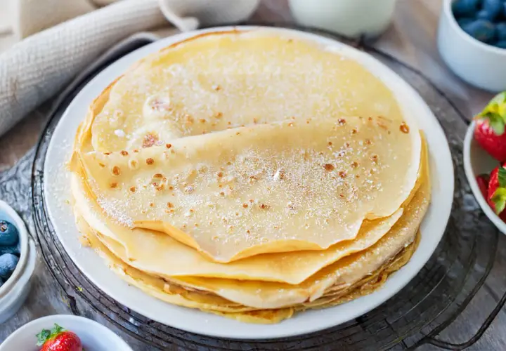 Ein Stapel goldbrauner Pfannkuchen, bestäubt mit Puderzucker. Daneben Schüsseln mit Blaubeeren und Erdbeeren. Ein sehr gutes Pfannkuchen Rezept für den Genuss.