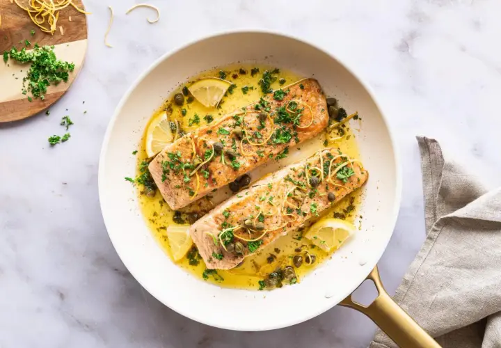 Zwei saftige Lachsfilets in einer Pfanne mit Zitronenscheiben, Kapern und Kräutern. Ein köstlicher Lachs mit Mangold-Aroma.
