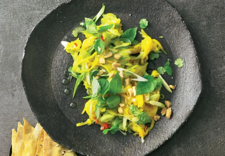 Ein bunter, frischer Mango Salat Thai mit Erdnüssen und Kräutern auf einem dunklen Teller.