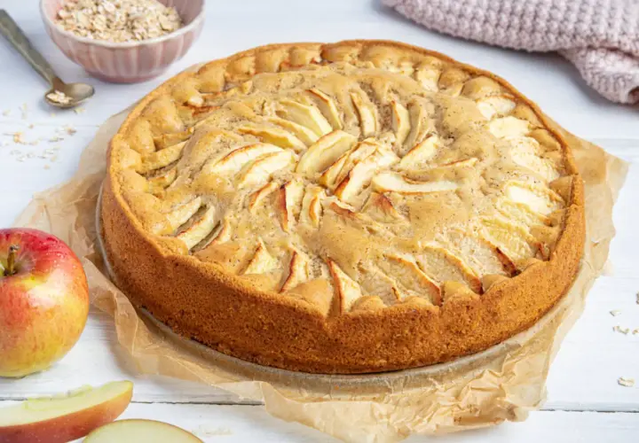 Ein goldbrauner, saftiger apfelkuchen ohne ei, dekoriert mit Apfelscheiben. Daneben liegen frische Äpfel und eine Schüssel Haferflocken.