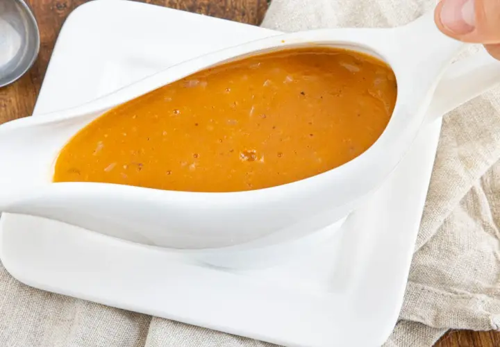 Eine köstliche, goldbraune Sauce mit Fond aus dem Glas, serviert in einer weißen Sauciere auf einem Leinentuch.