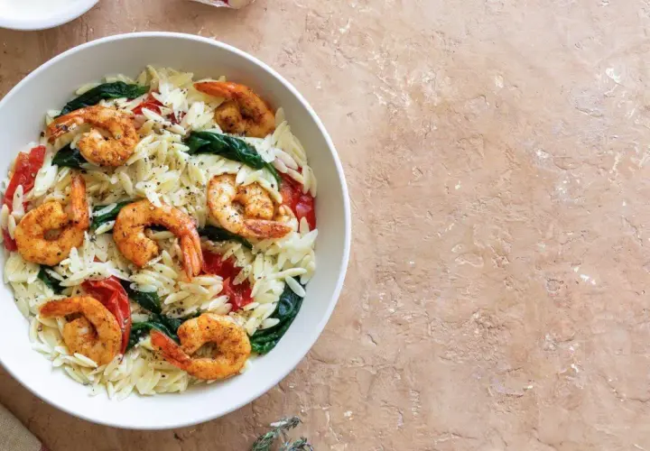 Ein Teller mit Orzo-Nudeln, Garnelen, Spinat und Tomaten, garniert mit Pfeffer. Dazu eine Zitronenspalte.