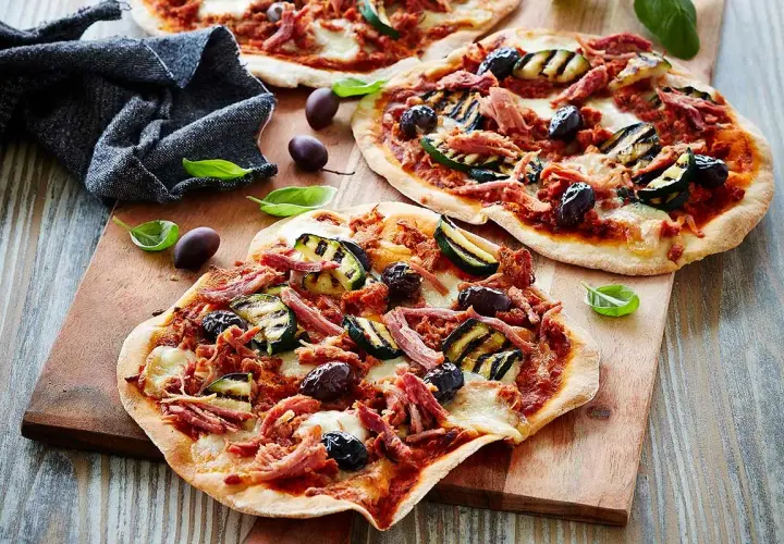 Drei kleine Pizzen mit pulled pork, gegrillter Zucchini und Oliven auf einem Holzbrett.