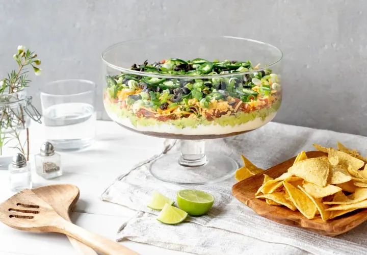 Schicht für Schicht ein Genuss: Ein bunter 7-Layer-Dip in einer Glasvitrine, bereit zum Dippen mit Tortilla-Chips.