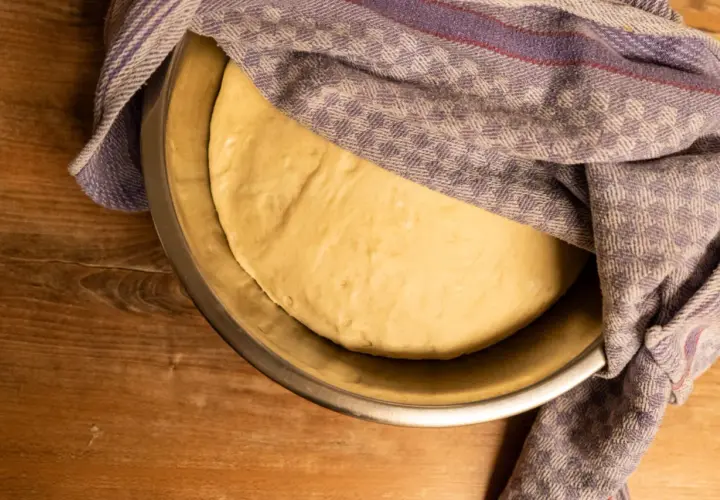 Frischer, aufgegangener Teig für pizzateig selber machen ruht in einer Schüssel unter einem karierten Tuch.