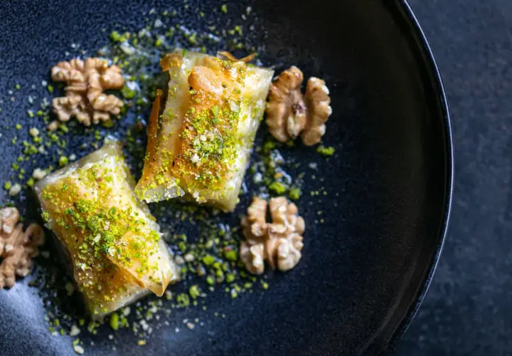 Zwei goldbraune Baklava-Stücke, bestreut mit gehackten Pistazien und Nüssen, auf einem dunklen Teller. Ein köstliches Baklava Rezept.