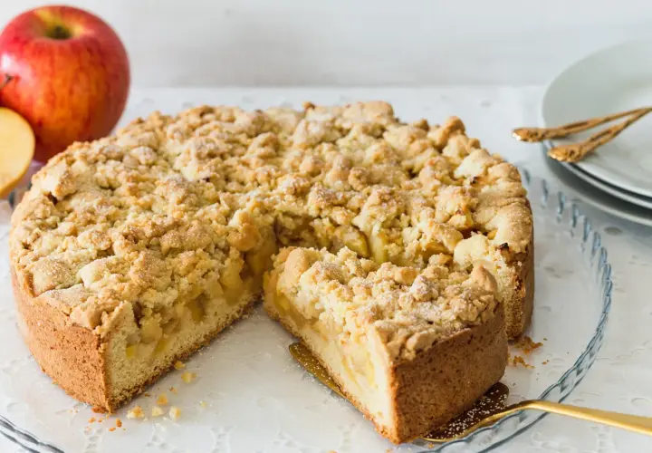 Ein saftiger, bester Apfelkuchen mit Streusel, bereit zum Servieren. Daneben liegen rote Äpfel.