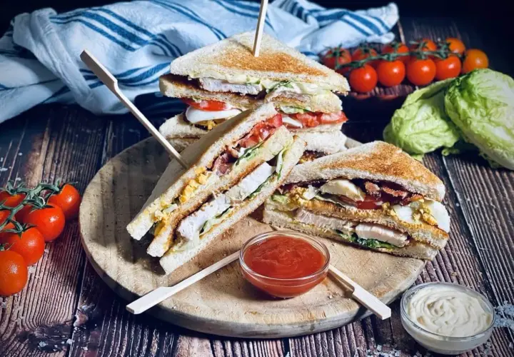 Drei saftige Club Sandwiches mit Hähnchen, Speck, Ei und Tomaten, serviert mit Ketchup und Mayo.