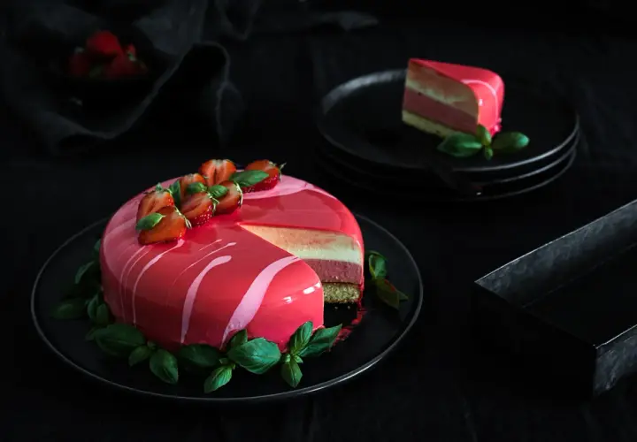 Eine Mousse Torte mit glänzendem Spiegelglasur, dekoriert mit Erdbeeren und Basilikum. Ein Stück ist auf einem Teller angerichtet.