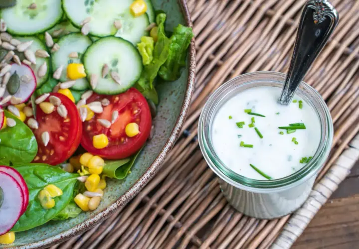Frischer Salat mit Tomaten, Gurken und Mais neben einem Glas mit selbstgemachtem Joghurt-Dressing, garniert mit Schnittlauch.