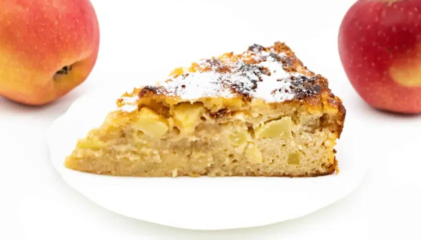 Ein Stück Apfelkuchen ohne Zucker mit Quark, bestäubt mit Puderzucker. Zwei Äpfel liegen daneben.