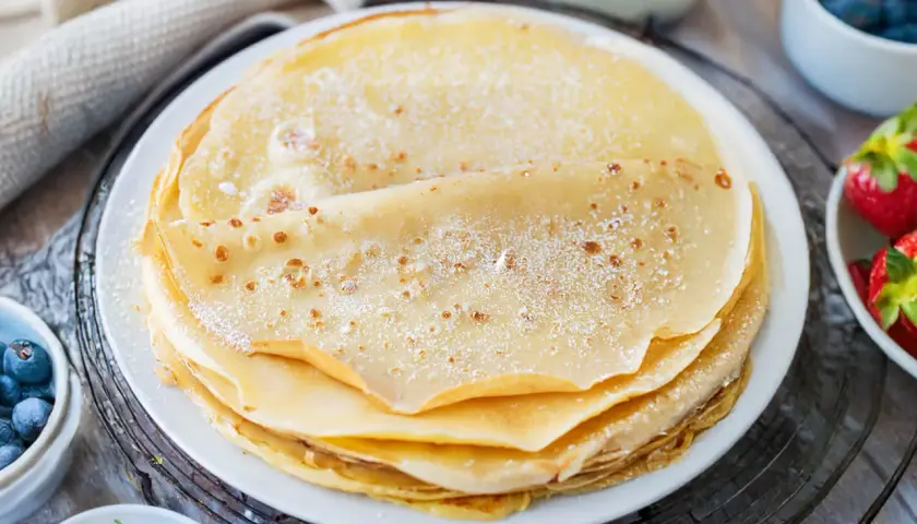Ein Stapel goldbrauner Pfannkuchen, bestäubt mit Puderzucker. Daneben Schüsseln mit Blaubeeren und Erdbeeren. Ein sehr gutes Pfannkuchen Rezept für den Genuss.