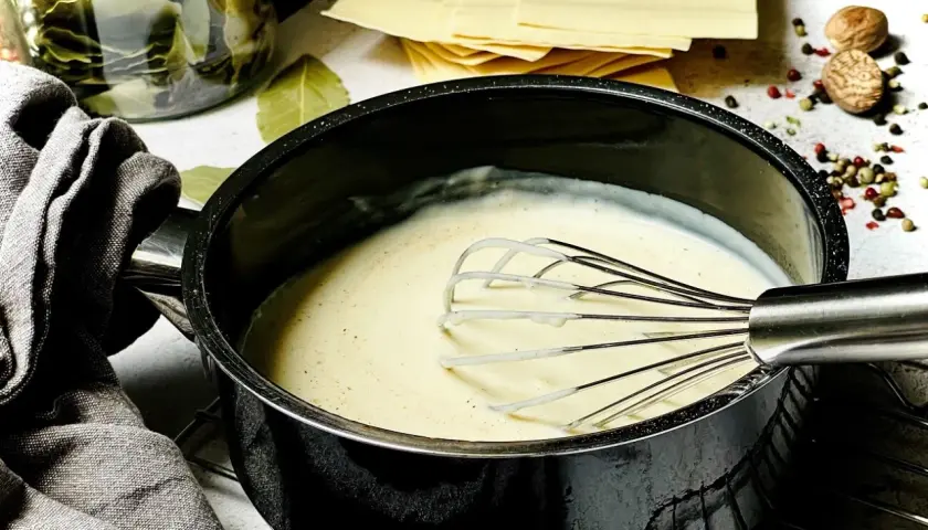 Cremige Béchamelsauce wird in einem Topf mit einem Schneebesen verrührt. Daneben liegen Lasagneplatten, Lorbeerblätter und Gewürze.