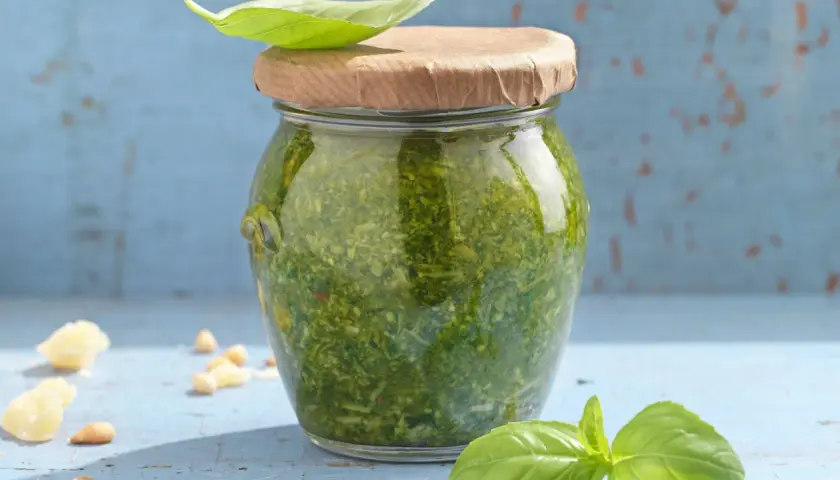 Frisches Pesto im Glas, garniert mit Basilikum. Zutaten wie Pinienkerne und Parmesan liegen daneben. Ein tolles pesto rezept!