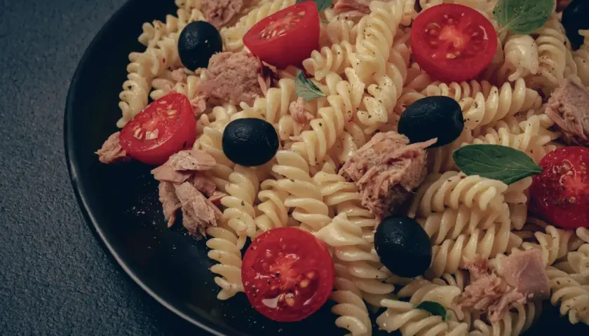 Ein köstlicher italienischer Nudelsalat mit Fusilli, Thunfisch, Kirschtomaten und schwarzen Oliven auf einem schwarzen Teller.