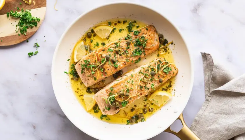 Zwei saftige Lachsfilets in einer Pfanne mit Zitronenscheiben, Kapern und Kräutern. Ein köstlicher Lachs mit Mangold-Aroma.