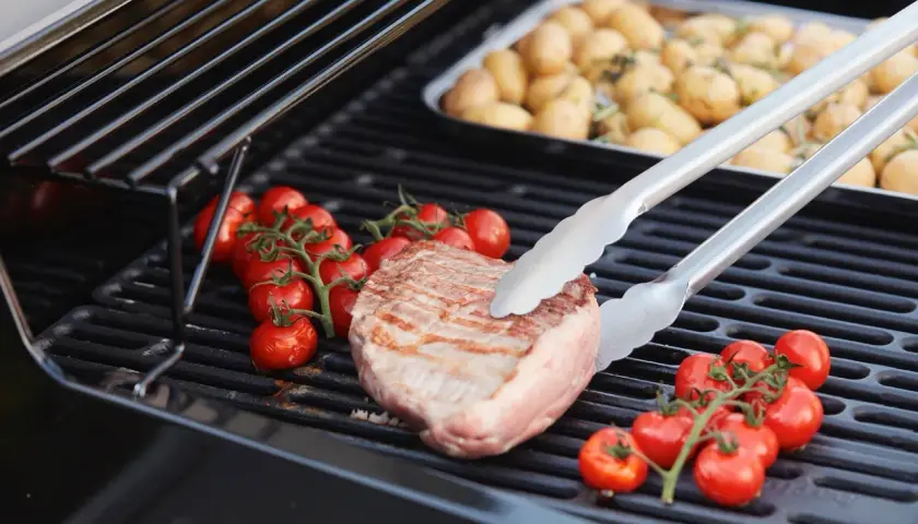 Duroc secreto zubereitung: Ein saftiges Steak wird mit Kirschtomaten auf dem Grill zubereitet. Daneben liegen Kartoffeln in einer Schale.