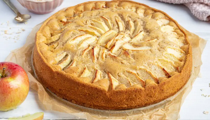 Ein goldbrauner, saftiger apfelkuchen ohne ei, dekoriert mit Apfelscheiben. Daneben liegen frische Äpfel und eine Schüssel Haferflocken.
