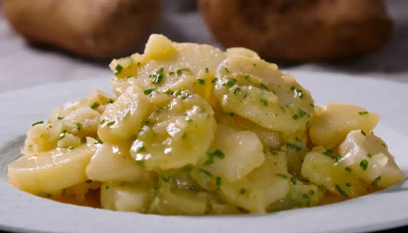 Ein Teller mit klassischem deutschen Kartoffelsalat, garniert mit frischen Kräutern. Im Hintergrund sind rohe Kartoffeln zu sehen.