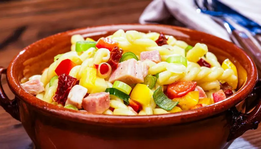Leckerer nudelsalat ohne mayonnaise mit bunten Paprikawürfeln, Schinken und Frühlingszwiebeln in einer Tonschüssel.
