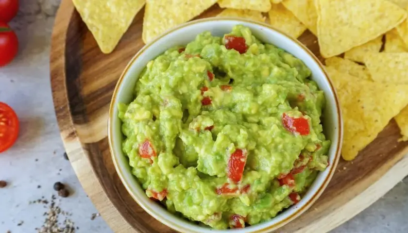 Frisches Guacamole-Rezept mit Tomatenstückchen, serviert mit Tortilla-Chips auf einem Holzbrett.