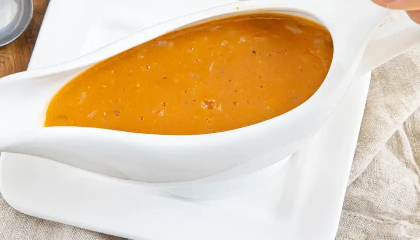 Eine köstliche, goldbraune Sauce mit Fond aus dem Glas, serviert in einer weißen Sauciere auf einem Leinentuch.