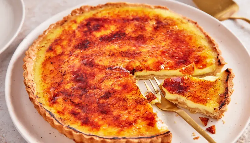 Eine cremige Crème brûlée Tarte mit karamellisierter Zuckerkruste, ein Stück wurde mit einer goldenen Gabel entnommen.