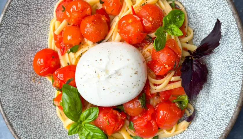 Ein cremiger Burrata Salat mit Pasta, Kirschtomaten und Basilikum auf einem gemusterten Teller.