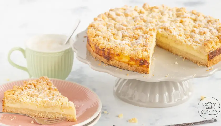 Ein saftiger Streuselkuchen mit Puddingfüllung, bestäubt mit Puderzucker, wartet auf Genießer. Daneben eine Tasse Cappuccino.