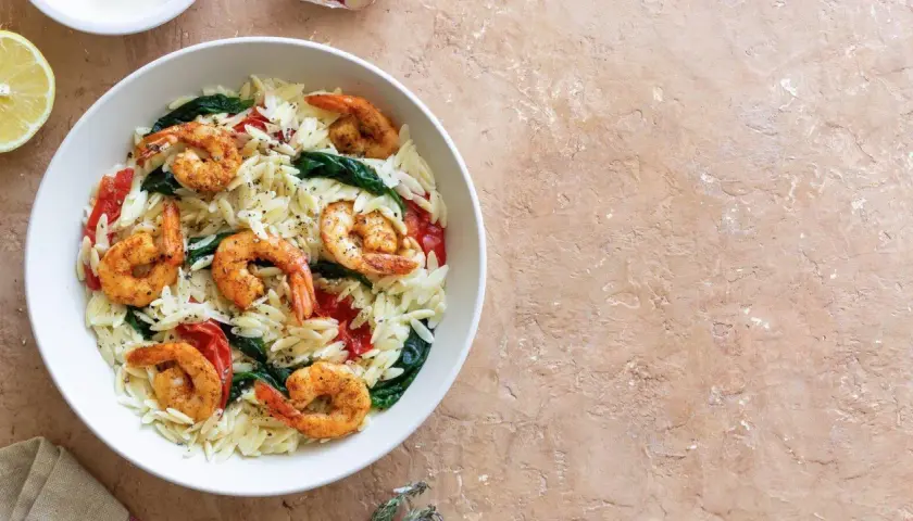 Ein Teller mit Orzo-Nudeln, Garnelen, Spinat und Tomaten, garniert mit Pfeffer. Dazu eine Zitronenspalte.
