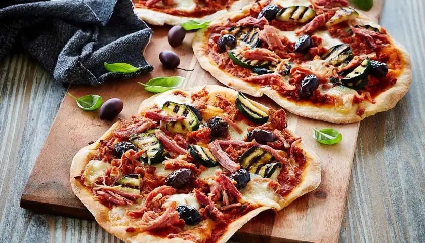 Drei kleine Pizzen mit pulled pork, gegrillter Zucchini und Oliven auf einem Holzbrett.